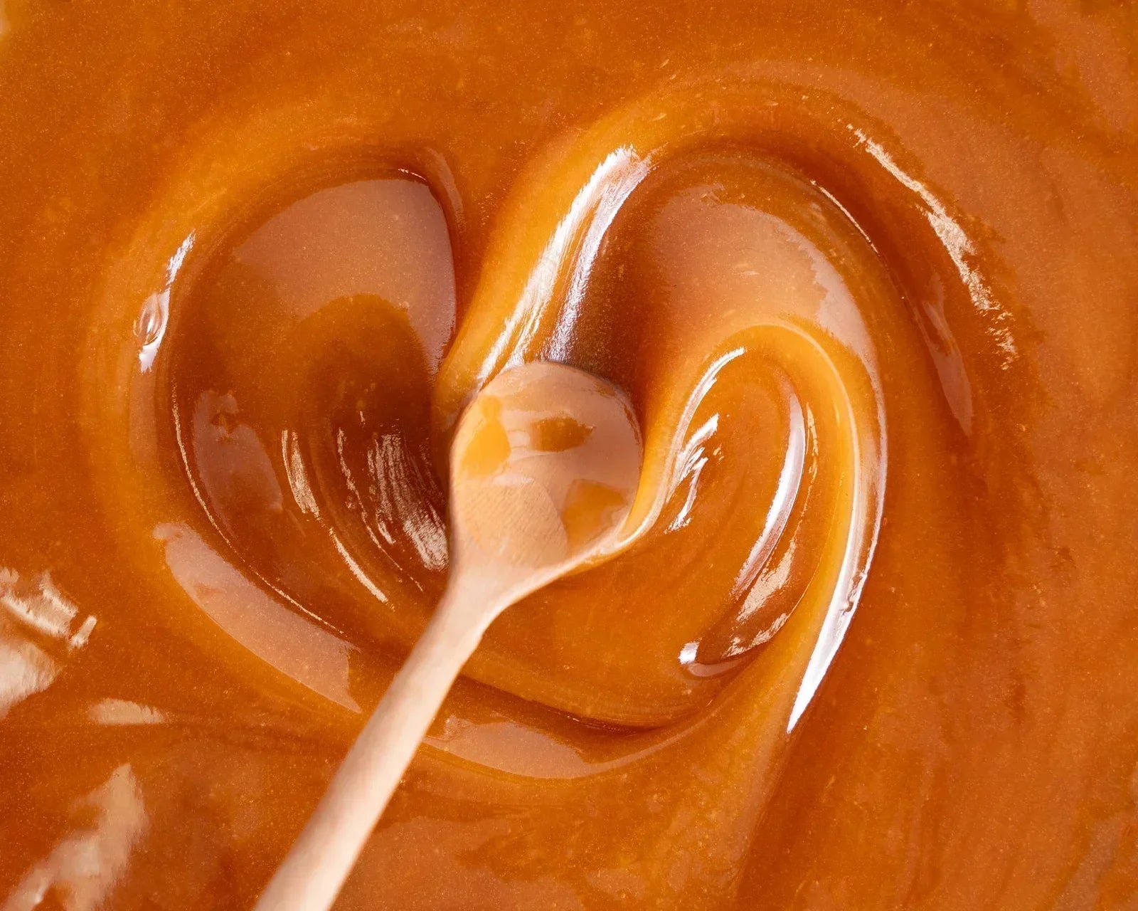 A wooden spoon stirring smooth, glossy caramel or toffee sauce, creating swirling patterns in the amber liquid.